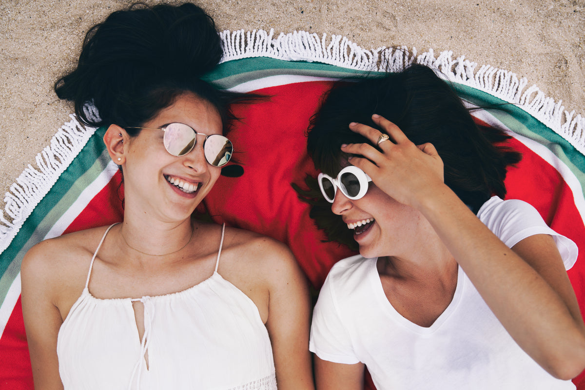 Two women laughing together on a round beach towel, relaxed inclusive summer lifestyle
